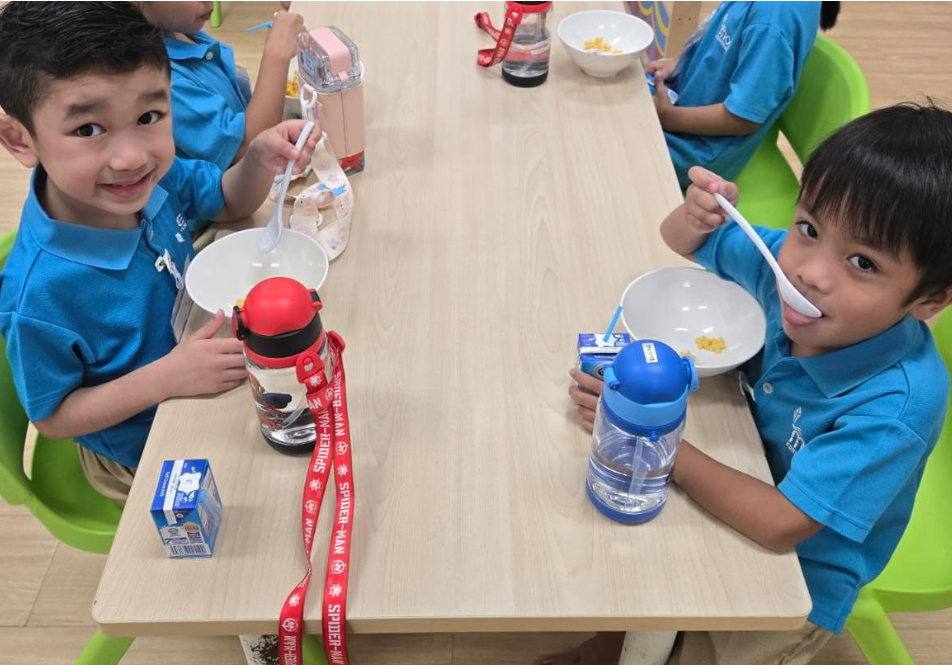 Two boys in blue uniforms sit at a table eating from bowls with spoons, water bottles and milk cartons nearby.